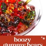 A glass bowl filled with colorful gummy bears and a gold spoon, labeled as “boozy gummy bears alcoholic treat.”.