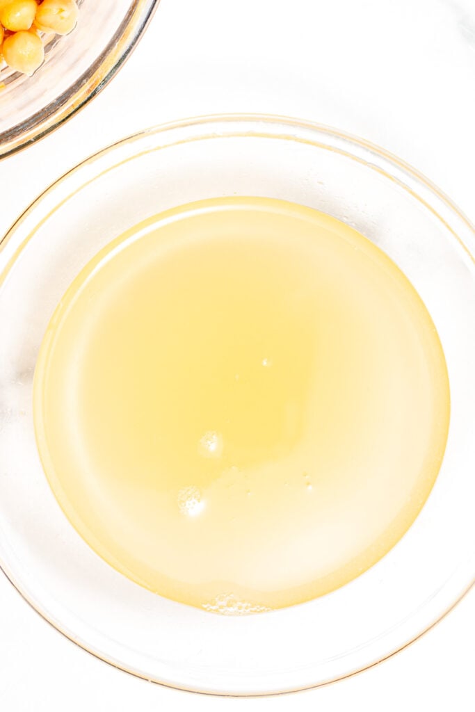 A clear glass bowl filled with light yellow liquid, with part of another bowl containing chickpeas visible in the corner.
