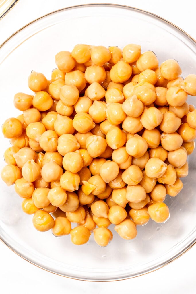 A clear glass bowl filled with cooked chickpeas on a white background.