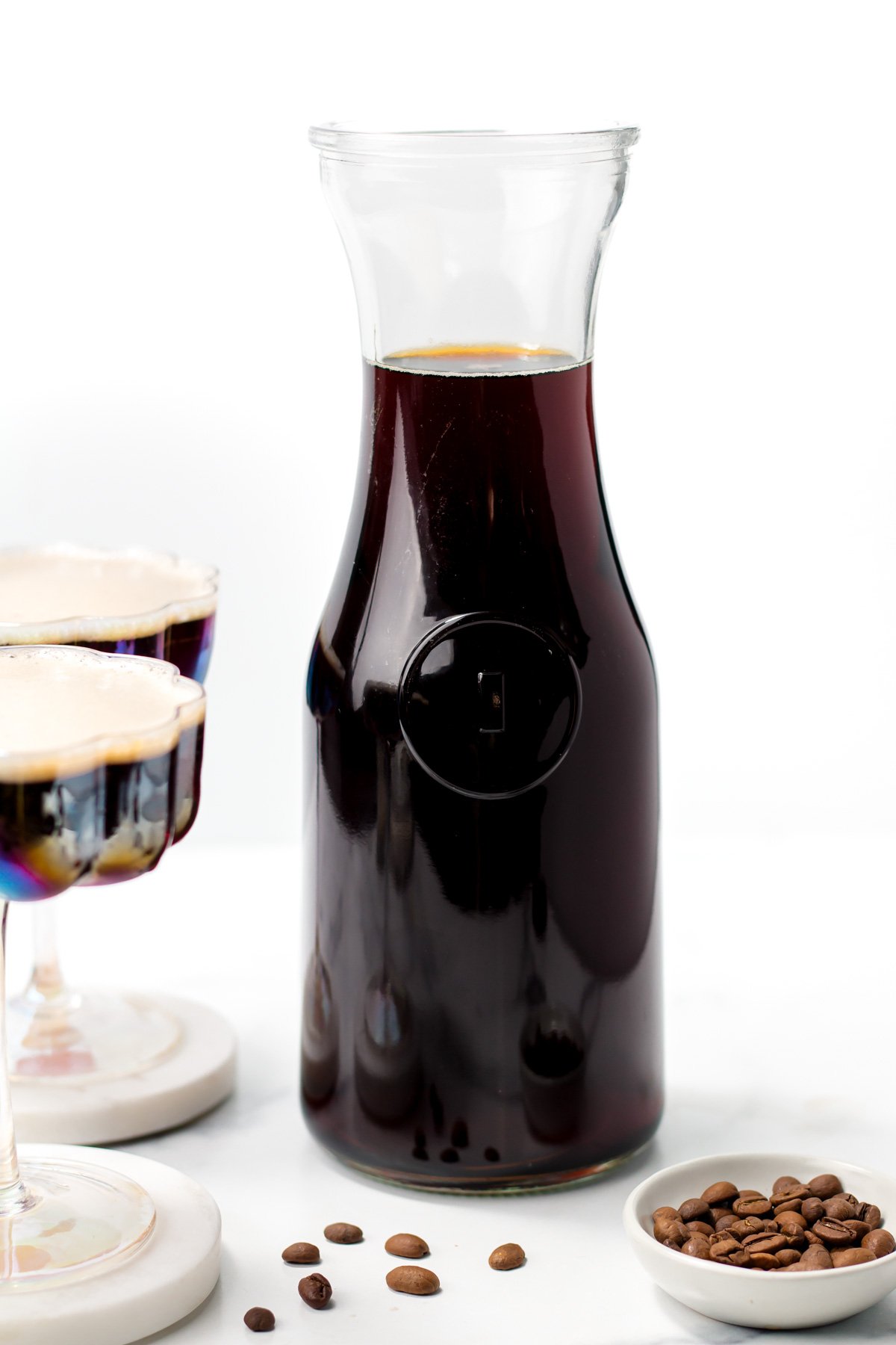A glass carafe filled with dark coffee, placed next to two glasses of coffee and a small dish of coffee beans on a white surface.