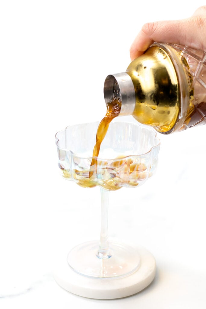 A hand pours a brown liquid from a gold cocktail shaker into a clear glass on a white surface.