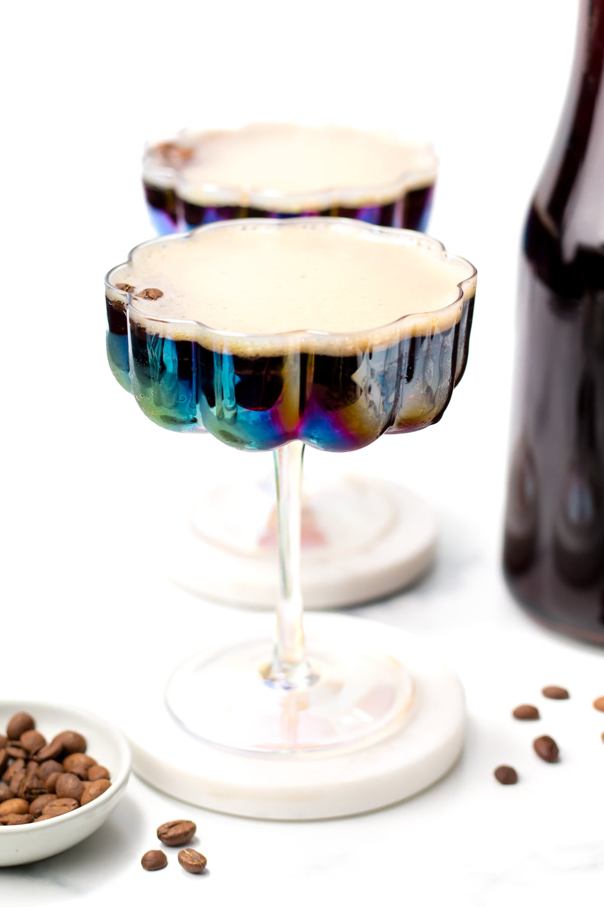Two coupe glasses filled with a dark creamy cocktail, possibly espresso martinis, sit on white coasters next to coffee beans and a dark bottle.