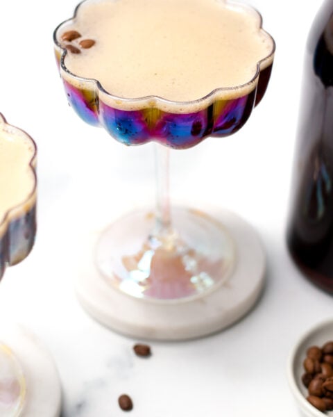 A frothy coffee cocktail in an iridescent glass sits on a marble coaster, with coffee beans scattered nearby and a bottle partially visible.