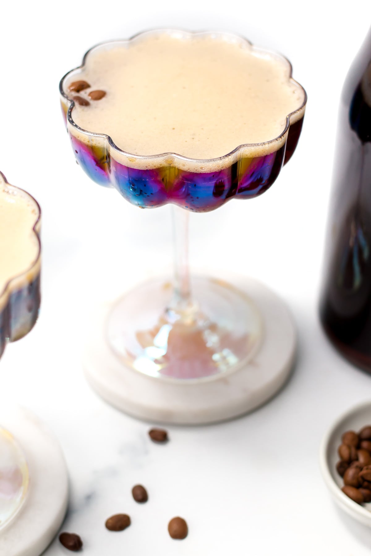 A frothy coffee cocktail in an iridescent glass sits on a marble coaster, with coffee beans scattered nearby and a bottle partially visible.