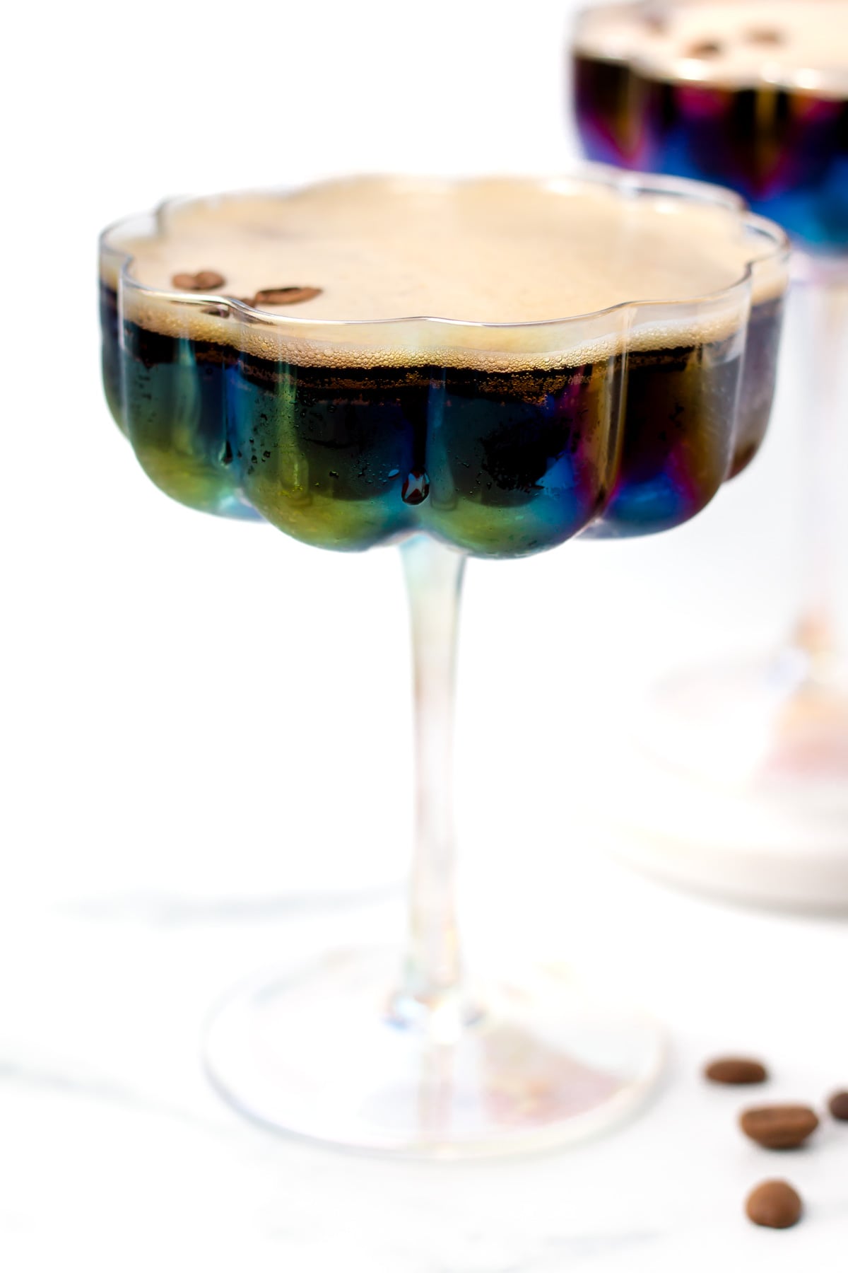 A glass with a scalloped edge filled with a dark coffee drink topped with foam, garnished with coffee beans, sits on a white surface.