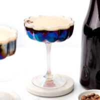 A glass of espresso martini sits on a coaster next to a bottle of dark liquid and scattered coffee beans.