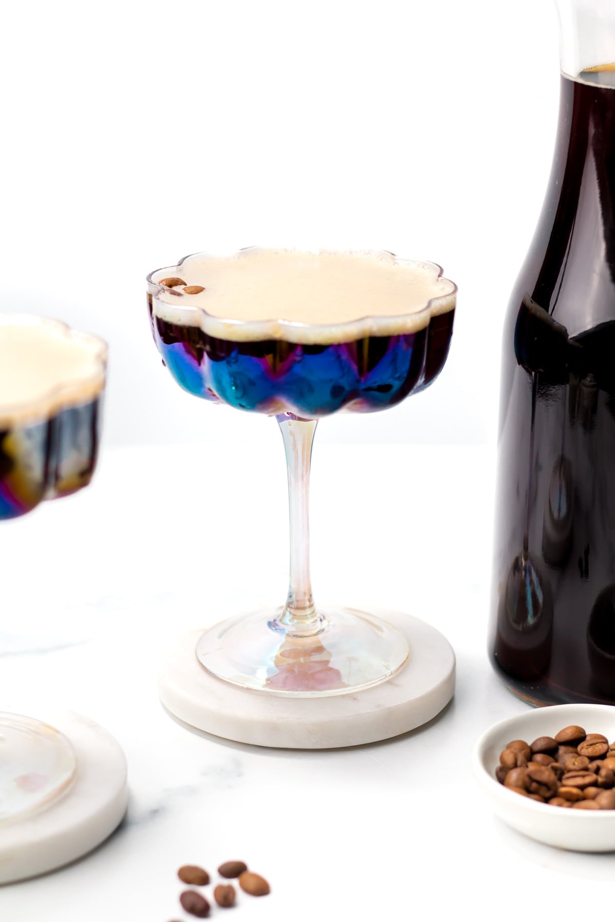 A glass of espresso martini sits on a coaster next to a bottle of dark liquid and scattered coffee beans.