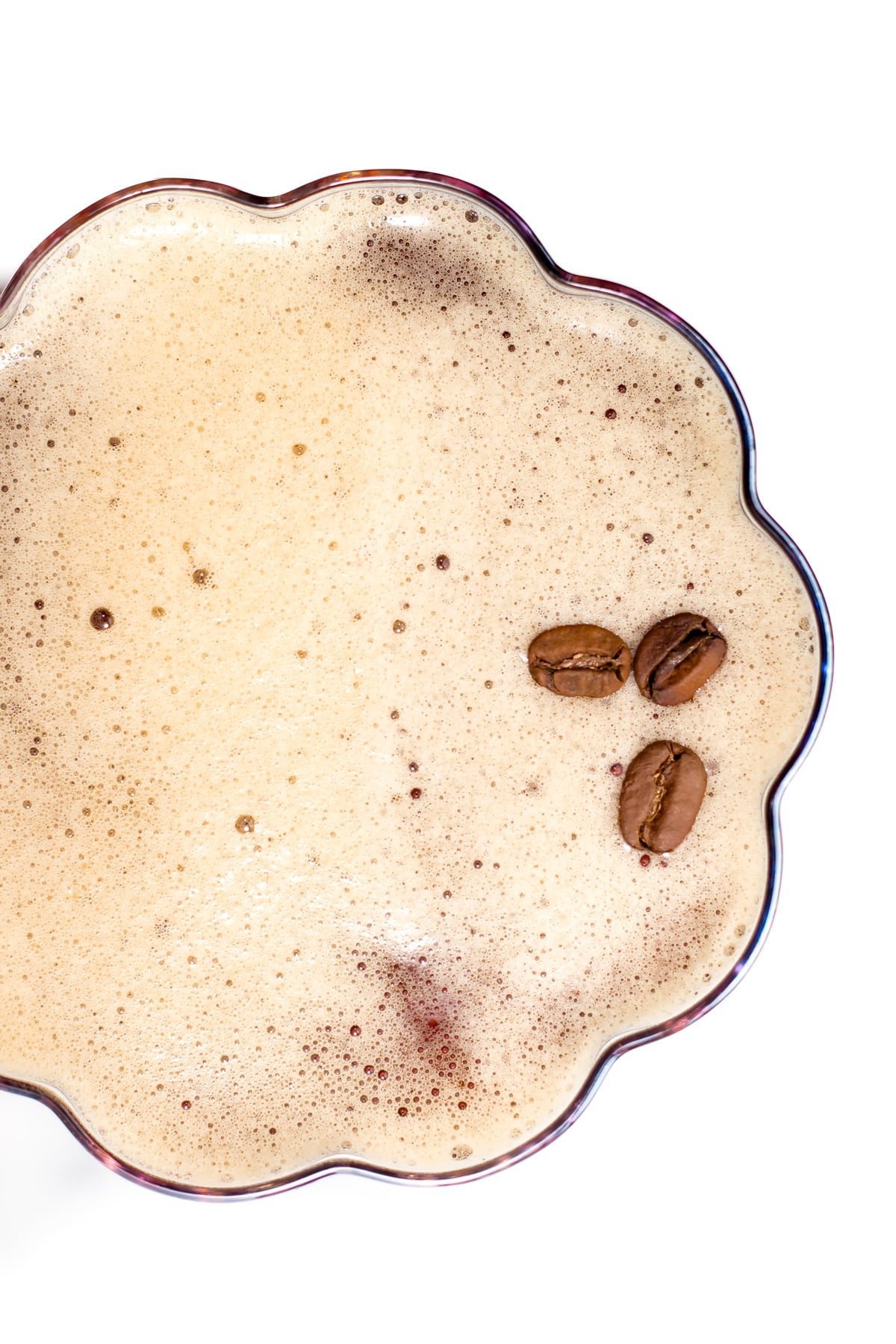 Top view of a frothy coffee drink in a scalloped-edged glass with three coffee beans on the foam, set against a white background.