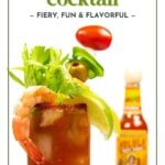 A Bloody Maria cocktail garnished with celery, shrimp, lime, olives, and cherry tomato, with a bottle of hot sauce in the background.