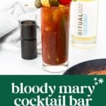 A Bloody Mary cocktail garnished with celery, pickles, olives, and a cherry tomato, sits next to a pepper grinder and a bottle of Ritual non-alcoholic spirit.