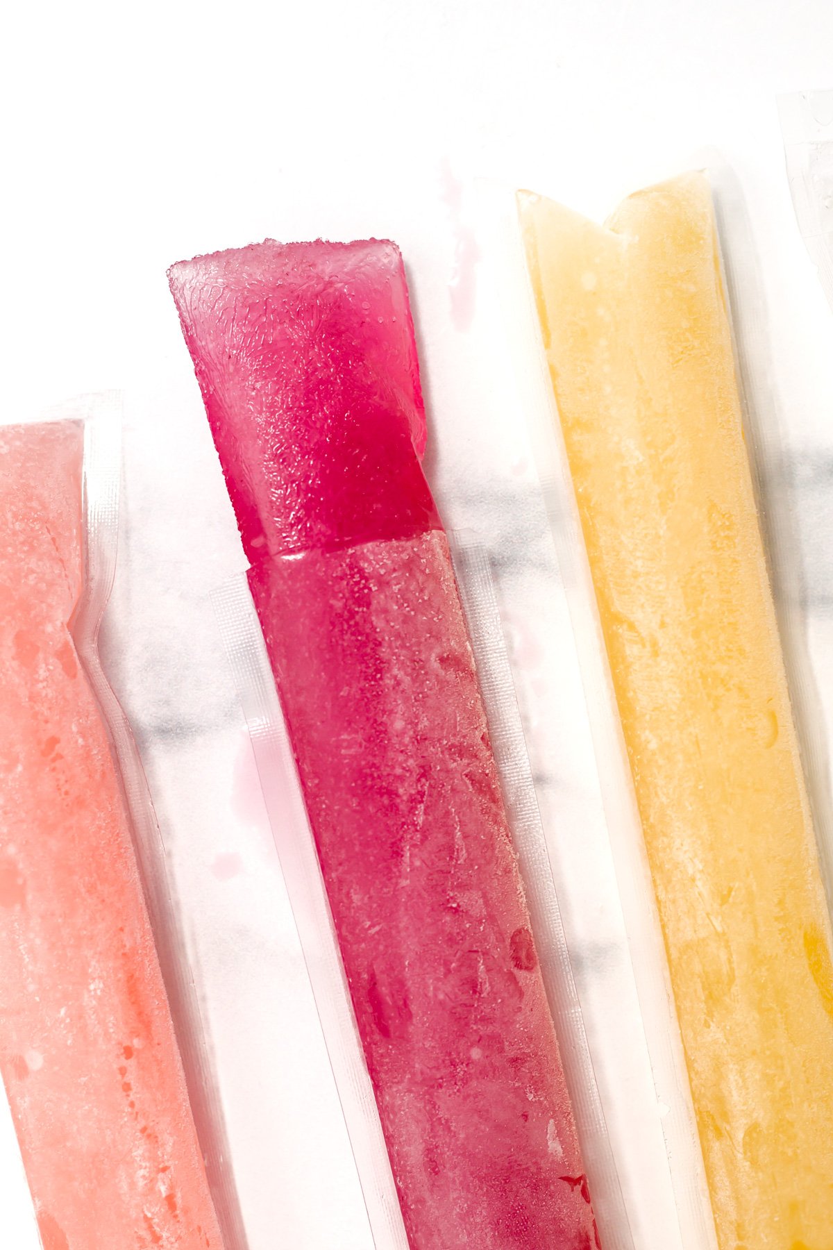 Three freeze pops in pink, red, and yellow colors are shown in clear plastic sleeves on a white surface.