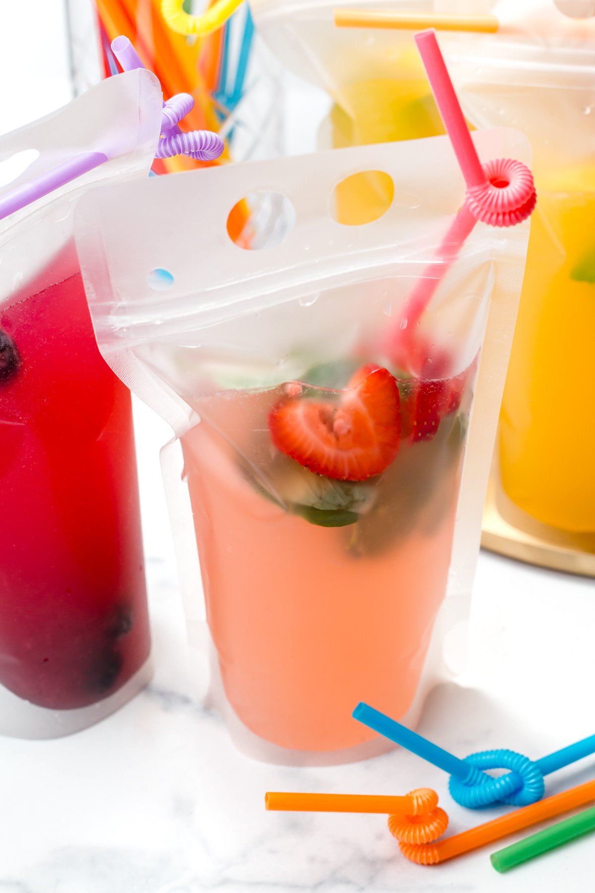 Clear drink pouches filled with colorful beverages, garnished with fruit and mint, each with a curly straw, set on a light surface.