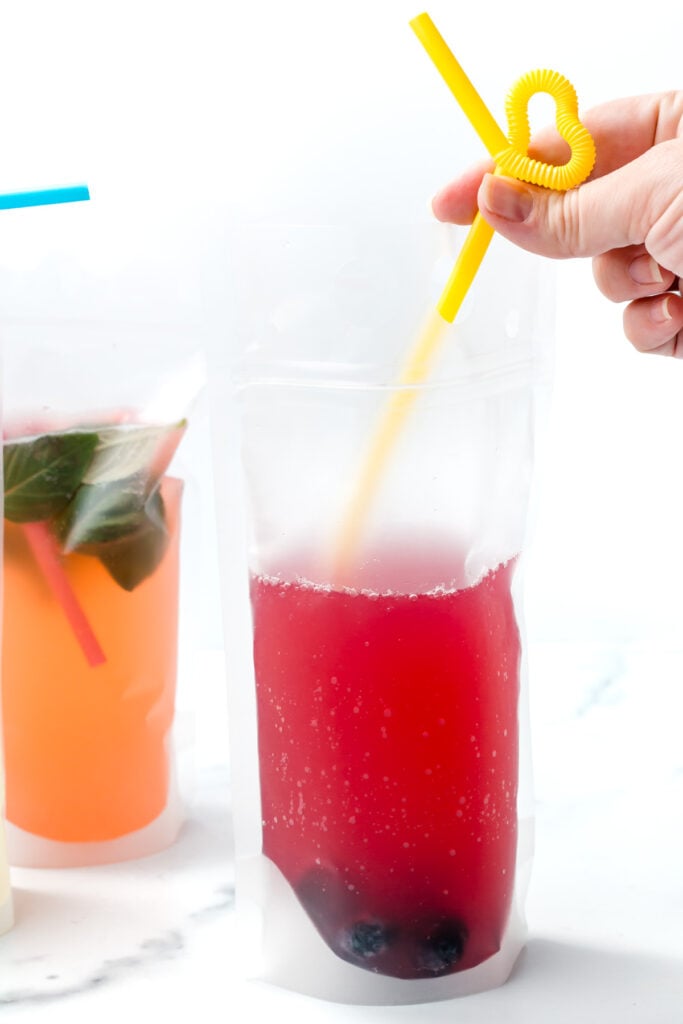 A clear plastic pouch of red juice with berries at the bottom and a yellow curly straw, next to another pouch with orange liquid and mint leaves.