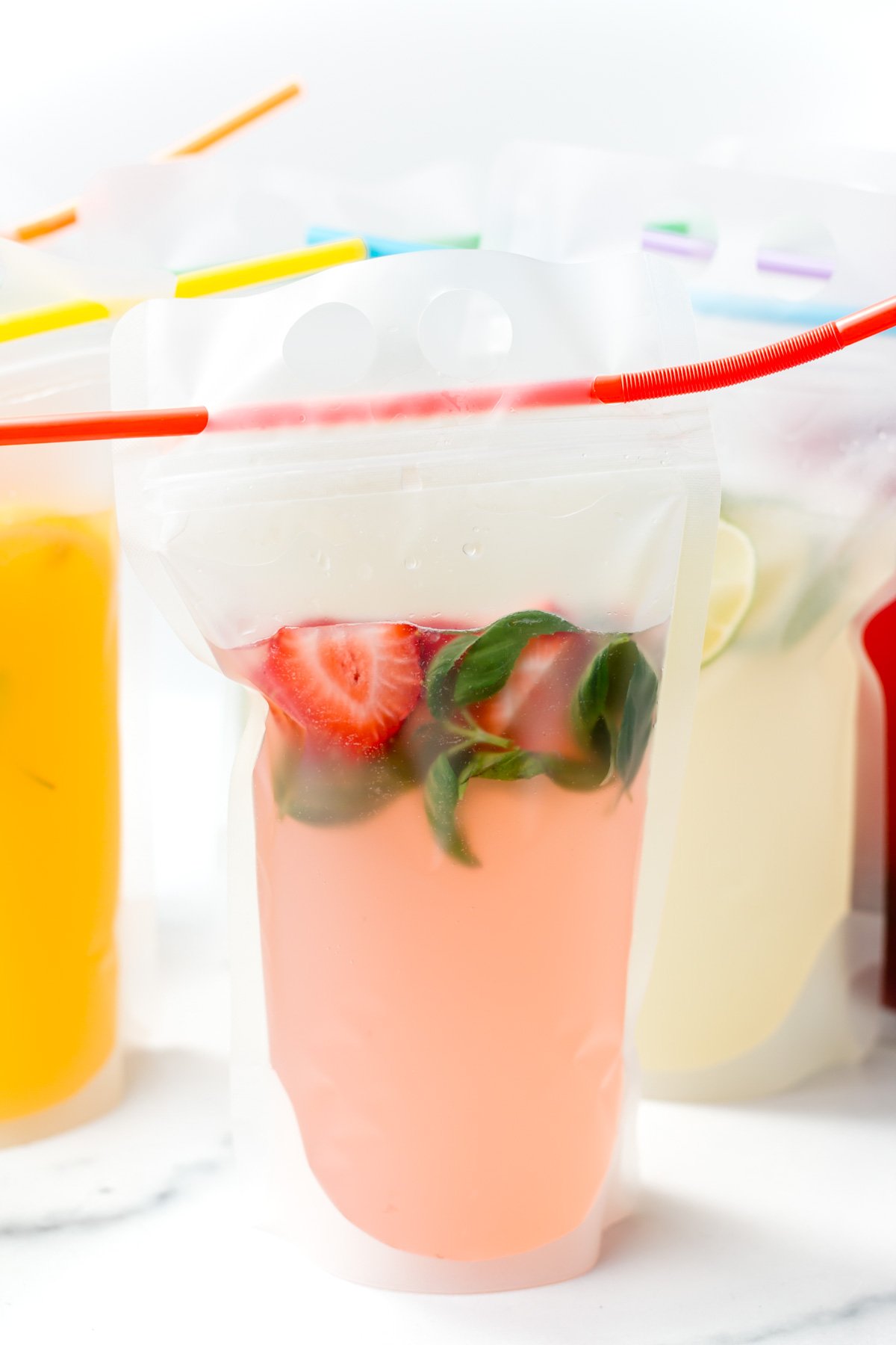 A clear drink pouch with pink beverage, fresh strawberries, basil leaves, and a red straw, with other colorful drink pouches in the background.