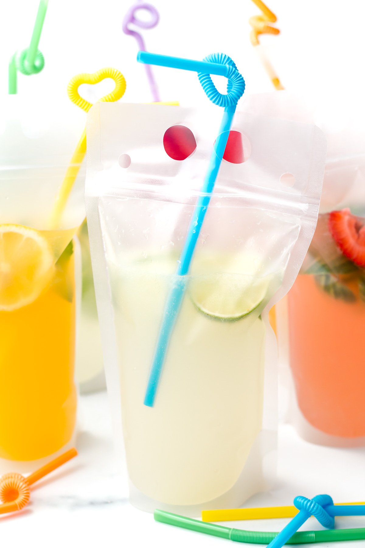 Clear plastic pouch of lemonade with lime slices and a blue curly straw, surrounded by other colorful drinks in similar pouches with decorative straws.