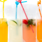 Four colorful drinks in clear plastic pouches with straws, featuring orange, yellow, and pink beverages, some with fruit and herbs inside.