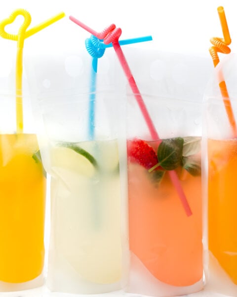 Four colorful drinks in clear plastic pouches with straws, featuring orange, yellow, and pink beverages, some with fruit and herbs inside.