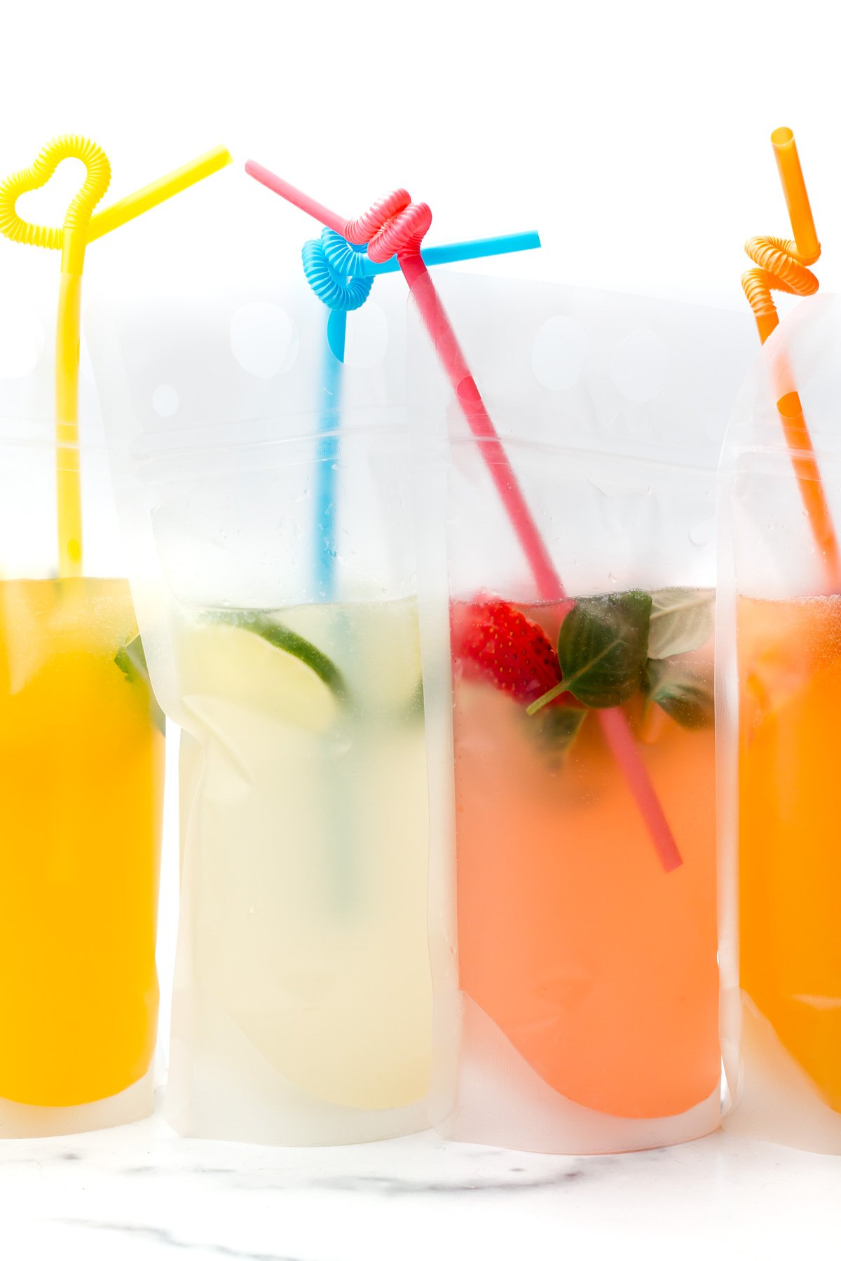Four colorful drinks in clear plastic pouches with straws, featuring orange, yellow, and pink beverages, some with fruit and herbs inside.