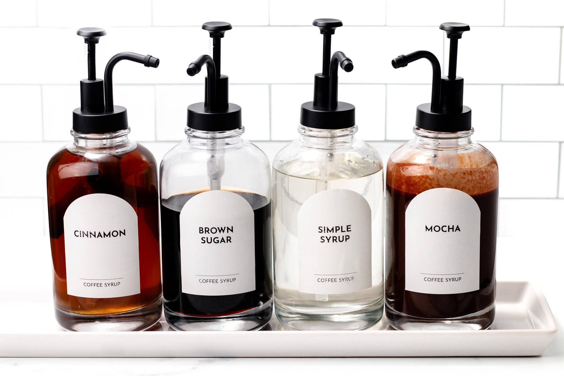 Four labeled pump bottles of coffee syrup—cinnamon, brown sugar, simple syrup, and mocha—are neatly arranged on a white tray against a tiled background, perfect for creating the ultimate coffee experience at your home Coffee Bar.
