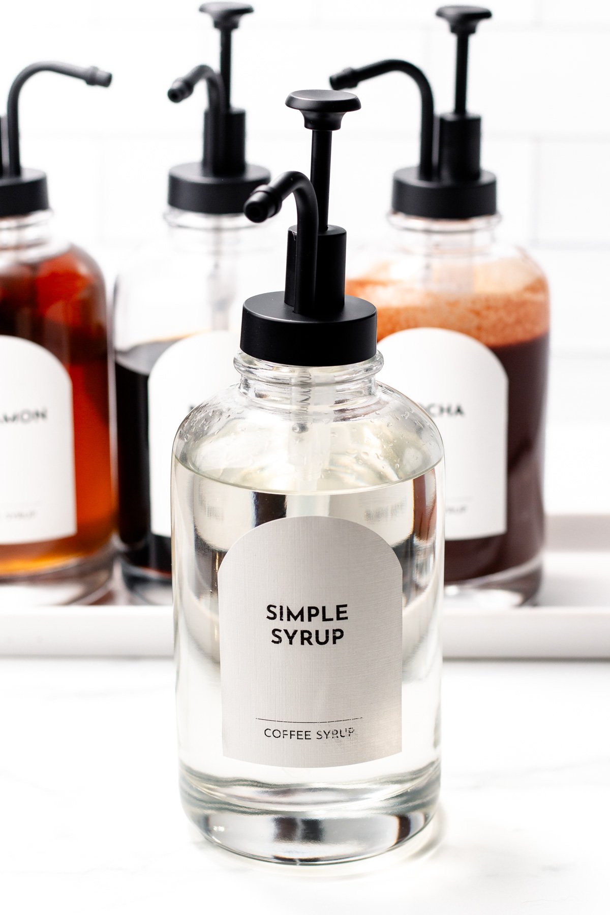 Clear glass pump bottle labeled "Simple Syrup" in front of three other syrup bottles, perfect for crafting the ultimate coffee at your home coffee bar.