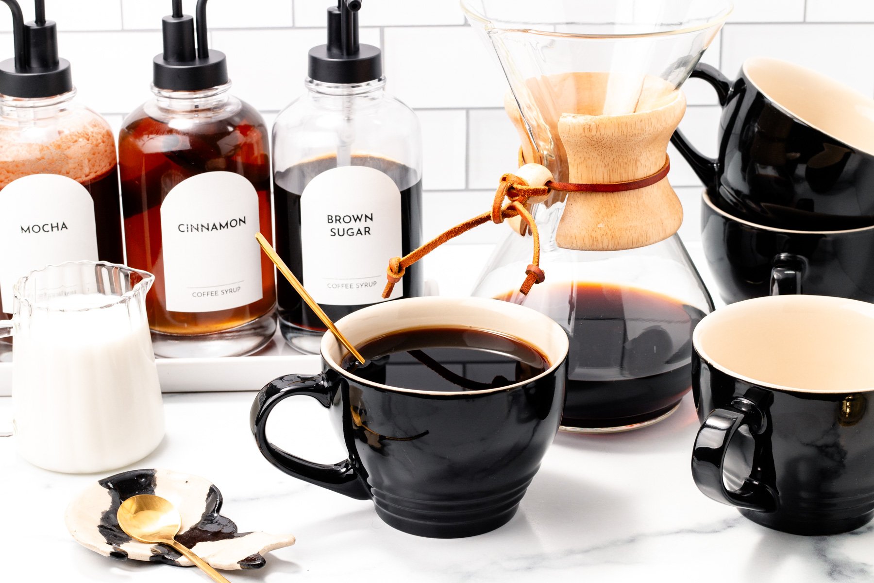 Three black coffee cups, a glass Chemex with coffee, coffee syrups labeled mocha, cinnamon, and brown sugar, and a small pitcher of milk create the perfect setup for an ultimate coffee bar on a white countertop.