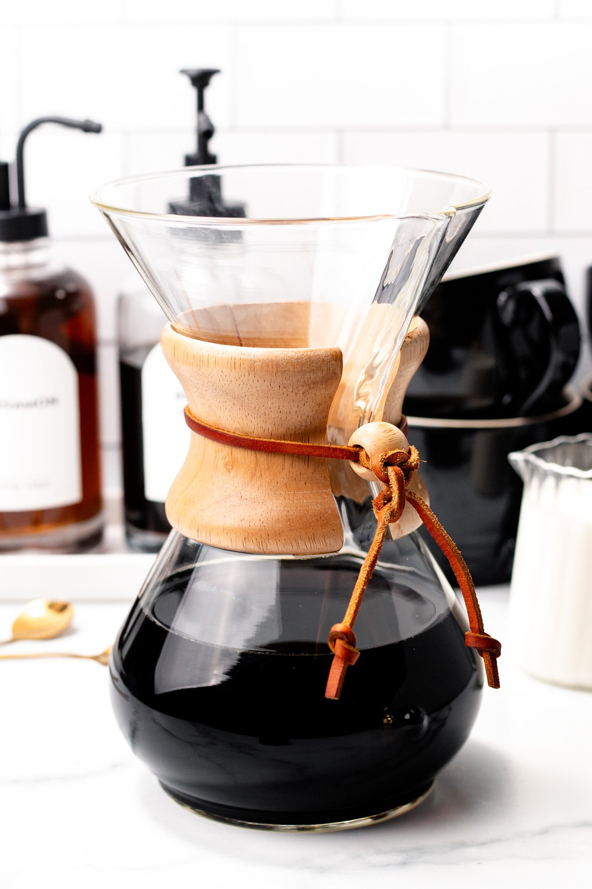 A glass Chemex coffee maker with a wooden collar and leather tie sits on a kitchen counter, ready to serve the ultimate coffee experience for your home coffee bar.