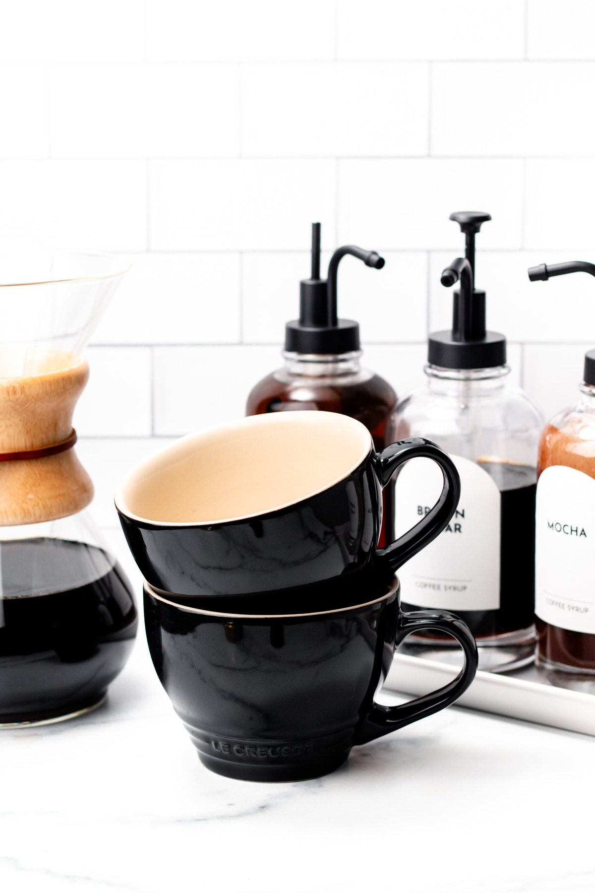 Two stacked black coffee mugs sit beside a coffee carafe and bottles of coffee syrup on a white countertop, creating the ultimate coffee bar against a tiled backsplash.