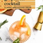 A glass of grapefruit cocktail with ice, garnished with a grapefruit slice and rosemary, sits on a white surface near a gold jigger and grapefruit slices.