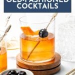 A glass of old-fashioned cocktail garnished with an orange peel and cherry, surrounded by a dish of cherries, cocktail picks, and a glass decanter on a white surface.