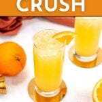A glass of orange-colored drink with ice and an orange slice garnish is on a coaster beside an orange and another filled glass. Text reads "Orange Crush" and "Get the recipe feastandwest.com.