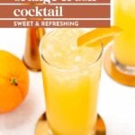 A tall glass of orange crush cocktail with ice and an orange slice garnish, next to a whole orange and a copper jigger on a white surface.