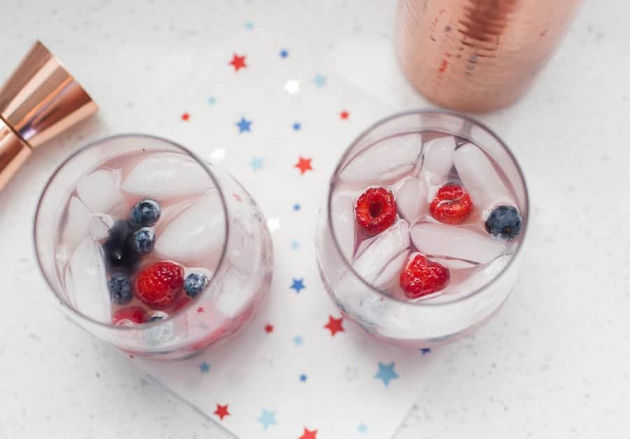 Two glasses with ice, raspberries, and blueberries on a star-patterned napkin, next to a copper cup and cocktail shaker.
