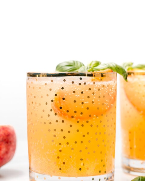 A glass of orange-colored sparkling beverage with ice, a peach slice, and basil garnish, served in a gold polka dot glass. Another similar glass and a peach are in the background.