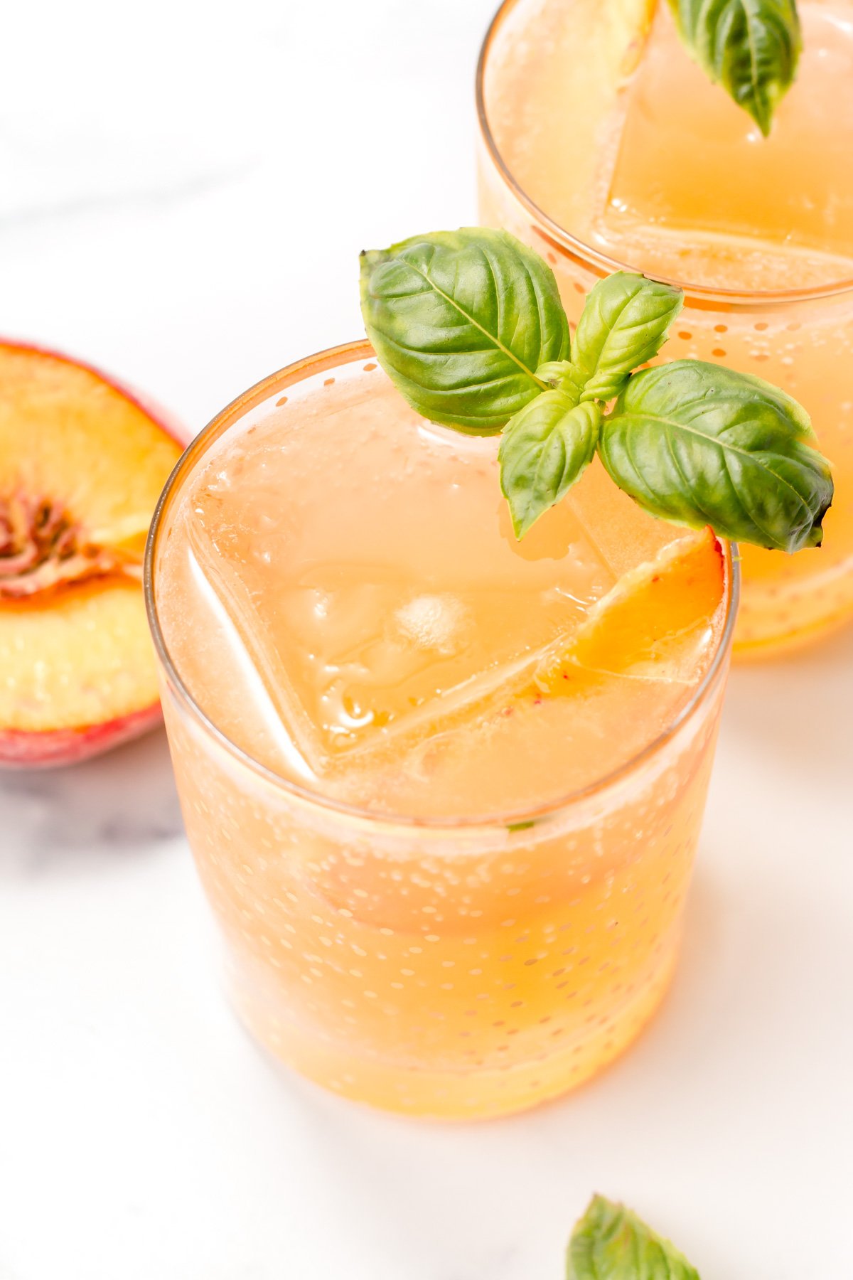 A glass of peach-colored sparkling drink with a large ice cube, garnished with fresh basil, next to a sliced peach on a white surface.