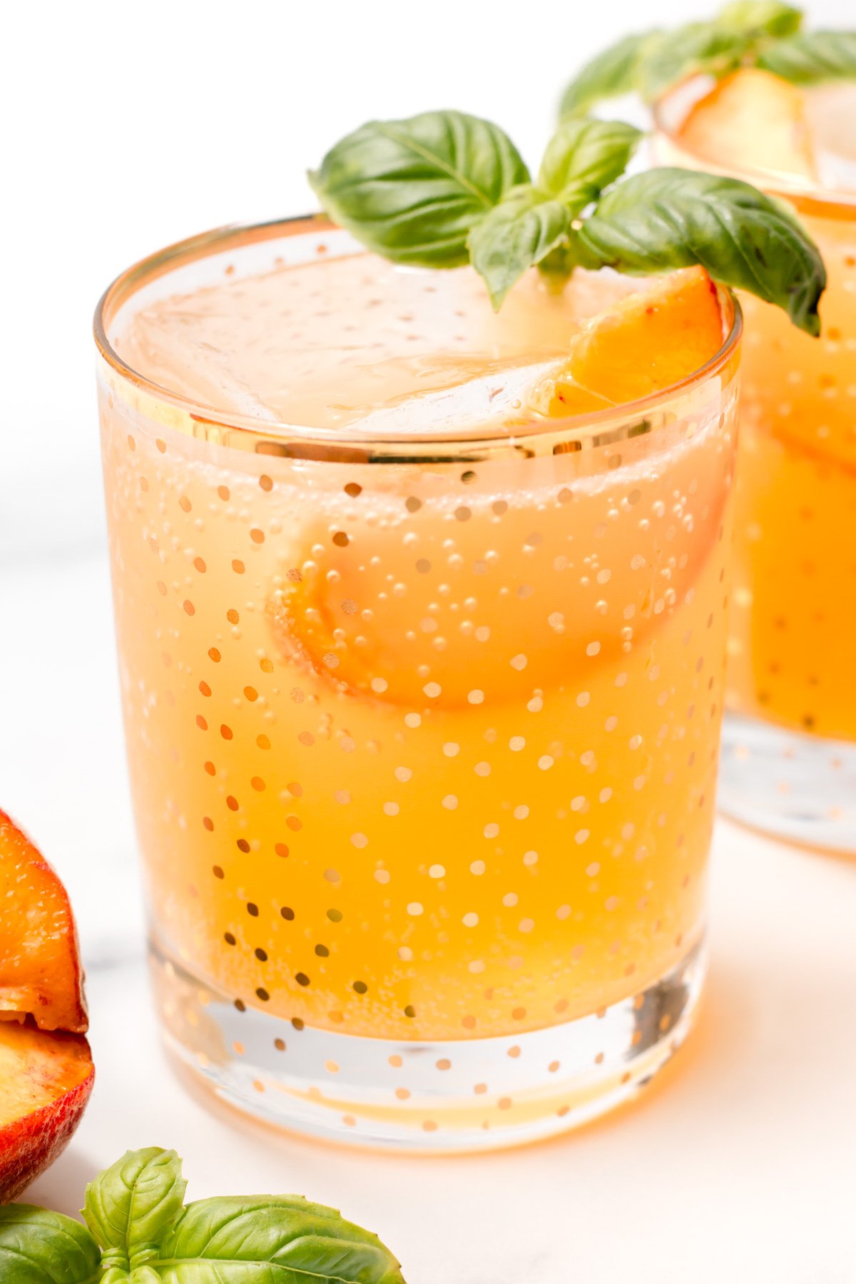 A glass filled with an orange-colored drink, garnished with peach slices and a sprig of basil, sits on a white surface.