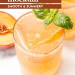 A glass of peach mocktail with ice, peach slice, and basil garnish sits next to a peach half on a white surface.