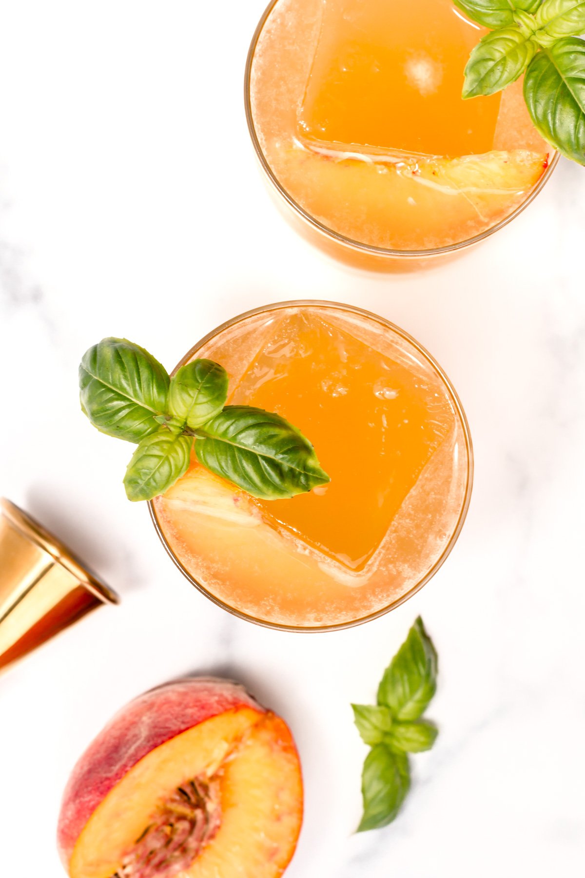 Two glasses of orange-colored drink with large ice cubes, garnished with basil leaves, next to a peach half and a gold jigger on a white surface.