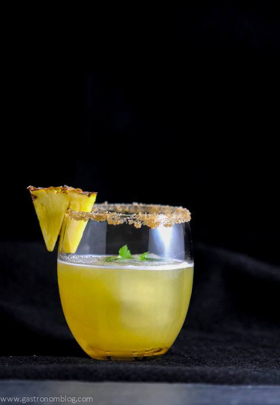 A yellow cocktail garnished with a pineapple wedge and brown sugar rim, served in a stemless glass against a black background.