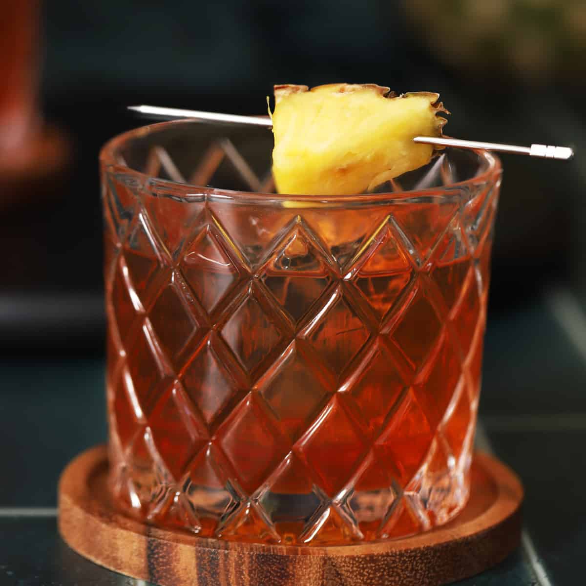 A glass of amber-colored cocktail on a wooden coaster, garnished with a pineapple wedge on a cocktail pick.