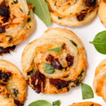 Close-up of savory pastry pinwheels filled with cheese, herbs, and sun-dried tomatoes, garnished with fresh basil leaves on a white background.