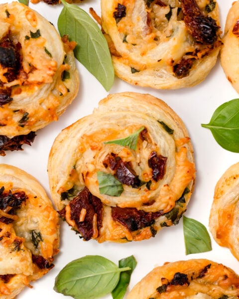 Close-up of savory pastry pinwheels filled with cheese, herbs, and sun-dried tomatoes, garnished with fresh basil leaves on a white background.