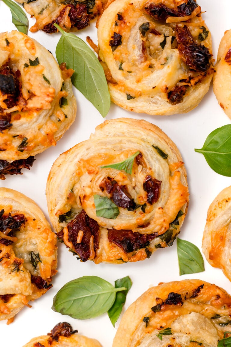 Close-up of savory pastry pinwheels filled with cheese, herbs, and sun-dried tomatoes, garnished with fresh basil leaves on a white background.