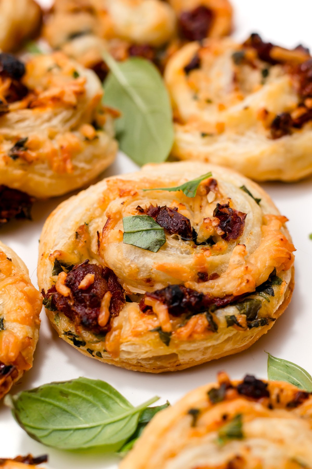 Close-up of savory puff pastry pinwheels filled with cheese, herbs, and sun-dried tomatoes, garnished with fresh basil leaves.