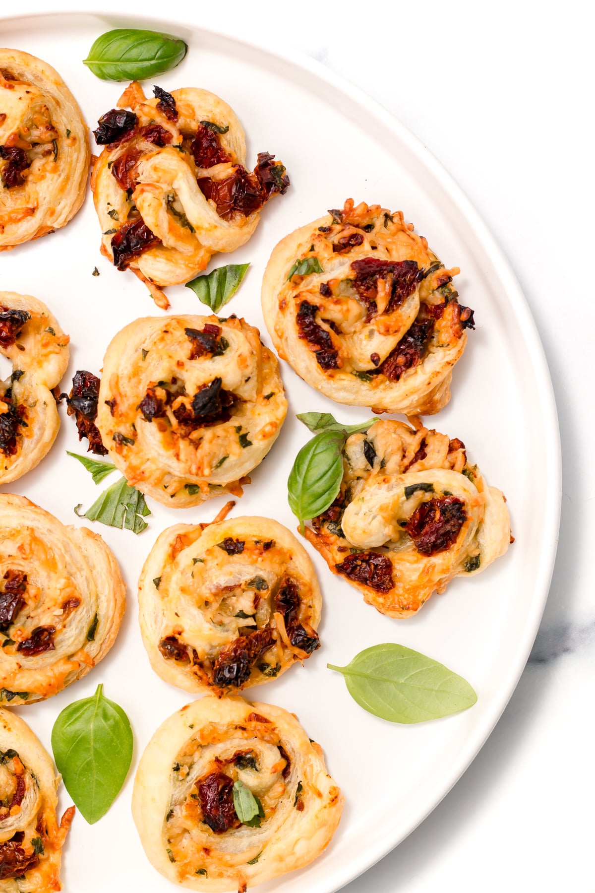A white plate with several puff pastry pinwheels filled with sun-dried tomatoes, cheese, and herbs, garnished with fresh basil leaves.