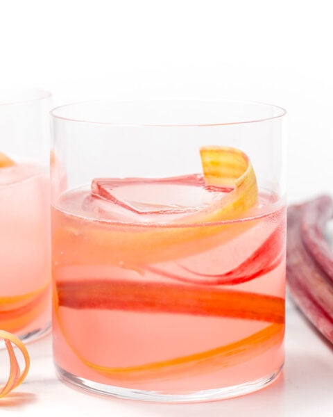 A clear glass filled with a pink beverage, ice cubes, and garnished with thin curly strips of rhubarb, placed on a white surface.