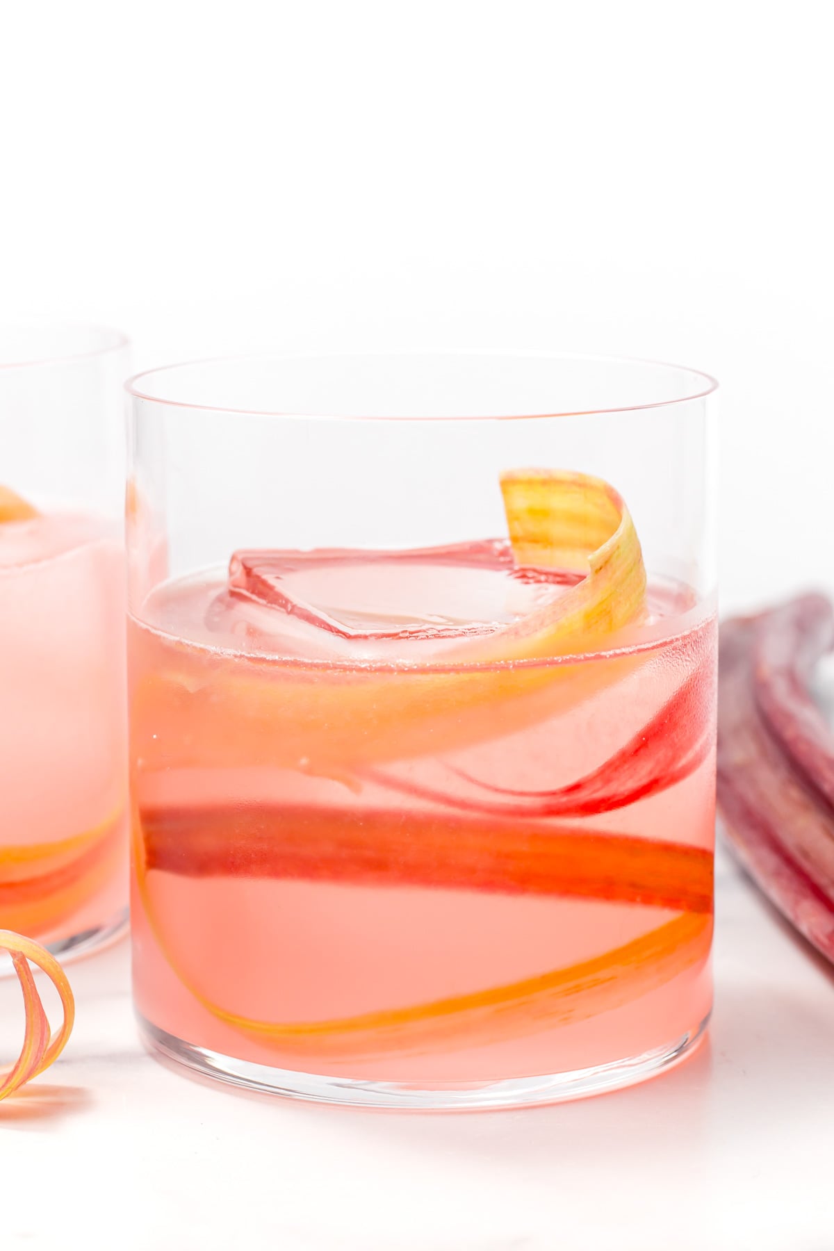 A glass of pink rhubarb cocktail with ice and rhubarb ribbons, set against a white background.