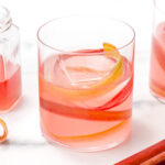 Three glasses of pink rhubarb cocktail with large ice cubes and rhubarb ribbons sit next to a jar of pink liquid on a white surface.