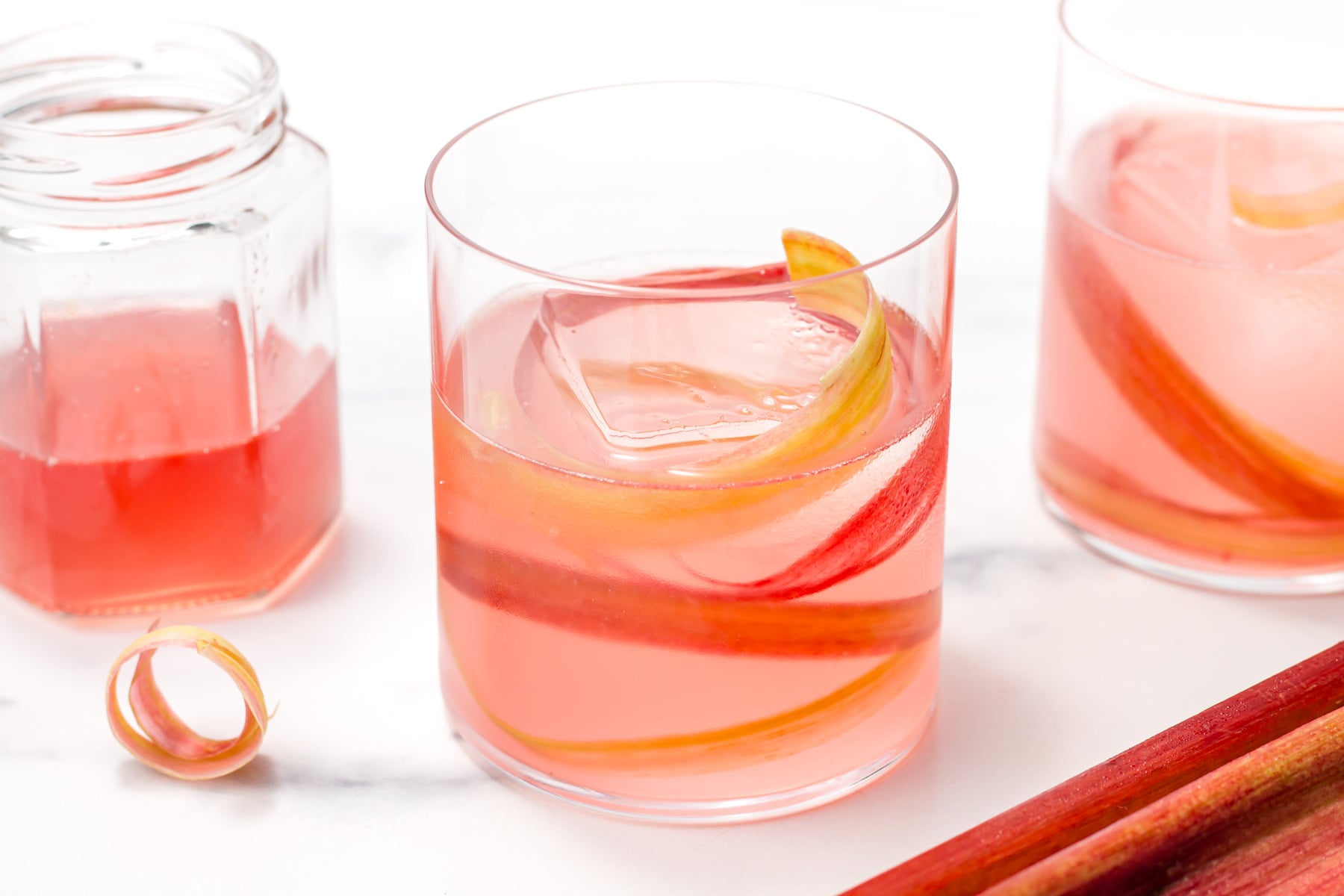 Two clear glasses of pink beverage with ice and rhubarb spiral garnish, next to a jar of pink liquid and a piece of rhubarb on a white surface.