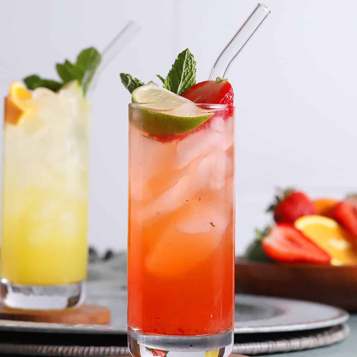 A tall glass of red iced drink garnished with lime, strawberry, and mint sits in front of a similar yellow drink; both have glass straws and are on a table with fruit in the background.