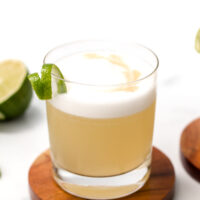 A glass of frothy cocktail garnished with a lime twist sits on a wooden coaster, with sliced limes and a knife nearby.